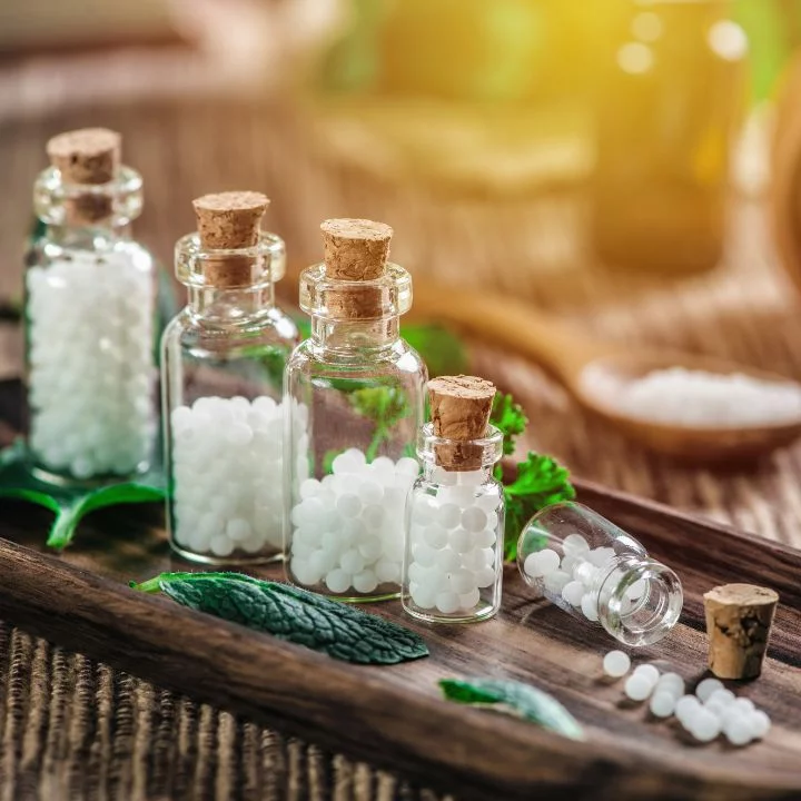 Kleine Apothekergläser mit weißen Homöopathie-Globuli auf einem Holztablett mit grünen Blättern